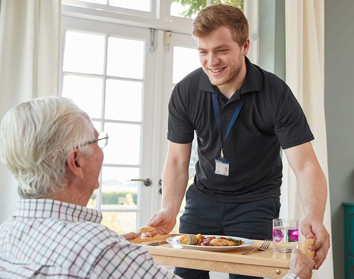 Eine Pflegekraft serviert einem älteren Herrn eine Mahlzeit auf dem Tablett